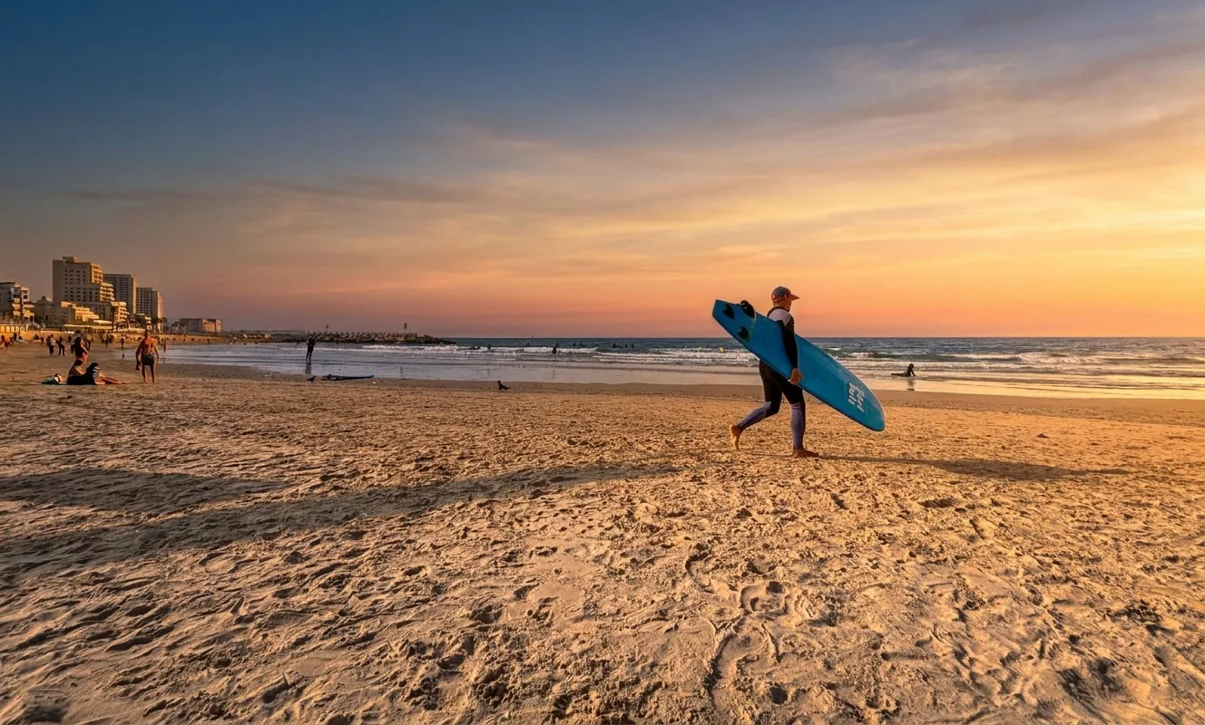 A surfer carrying a blue board along Tel Baruch beach, with a breakwater in the distance