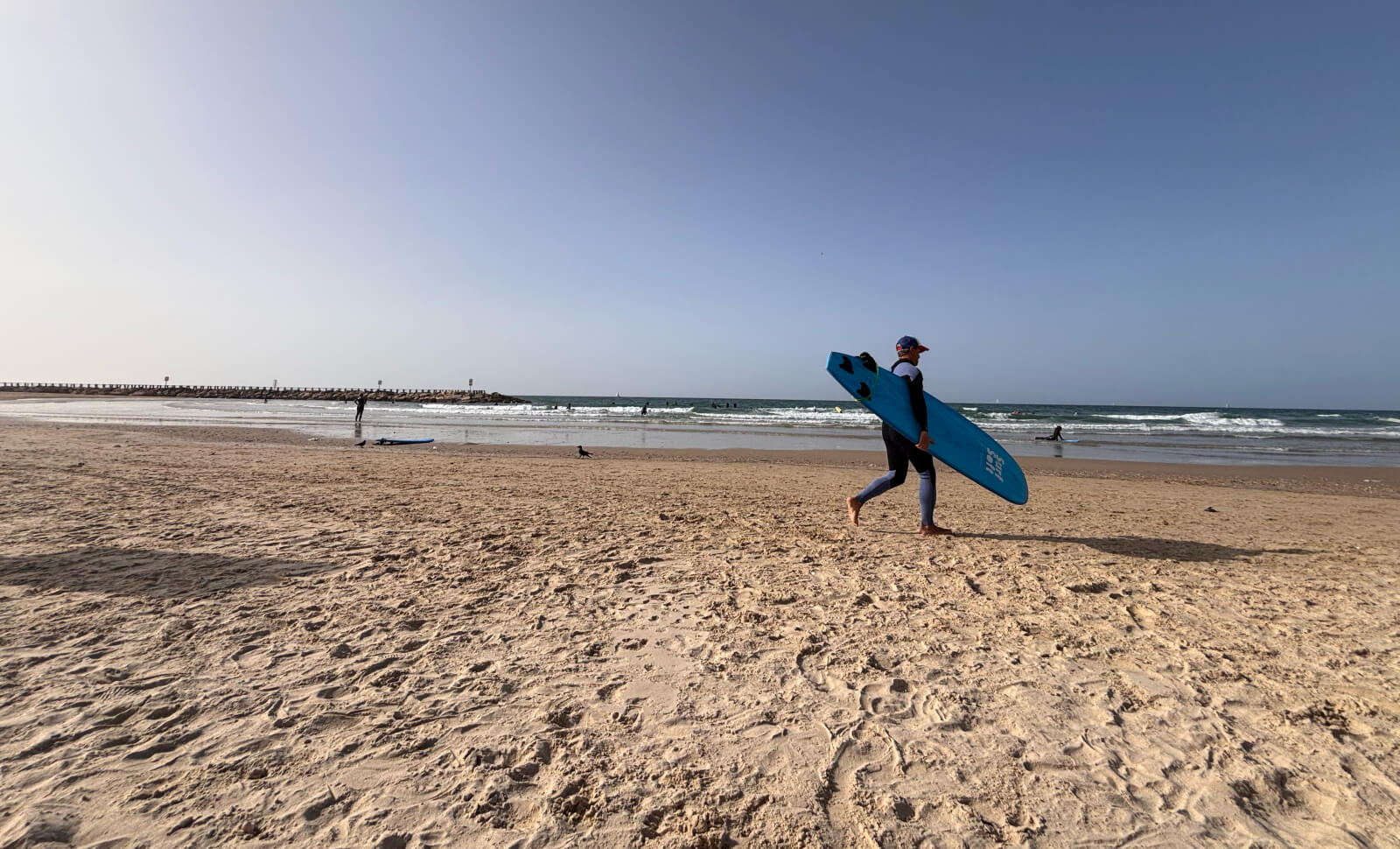 A surfer carrying a blue board along Tel Baruch beach, with a breakwater in the distance